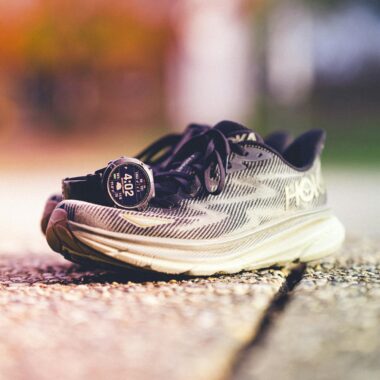 A pair of Hoka running shoes with a Garmin watch beside them on a sunny outdoor track.