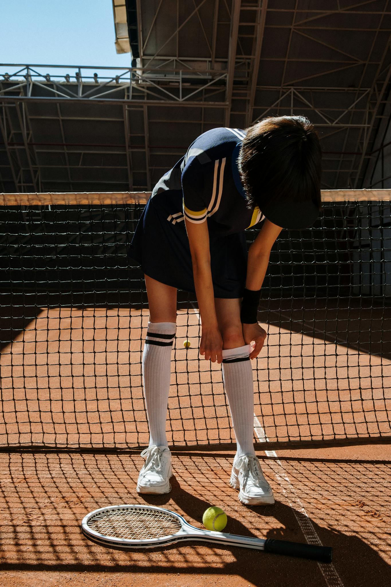 Tennis player adjusting gear on a clay court, with racket and ball on the ground.