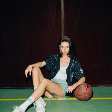 Young woman sitting with a basketball on a court, stylishly posing indoors.