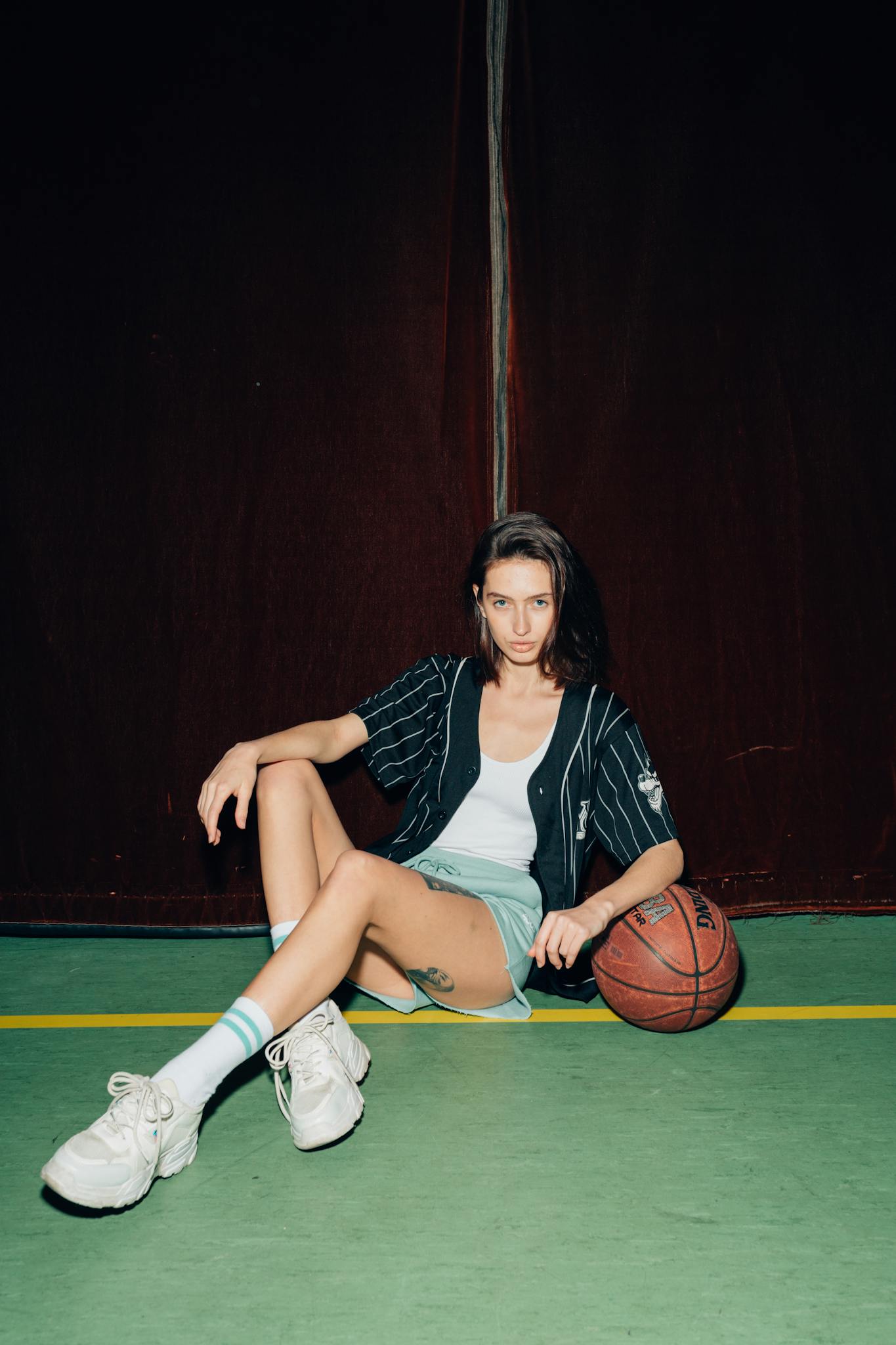 Young woman sitting with a basketball on a court, stylishly posing indoors.